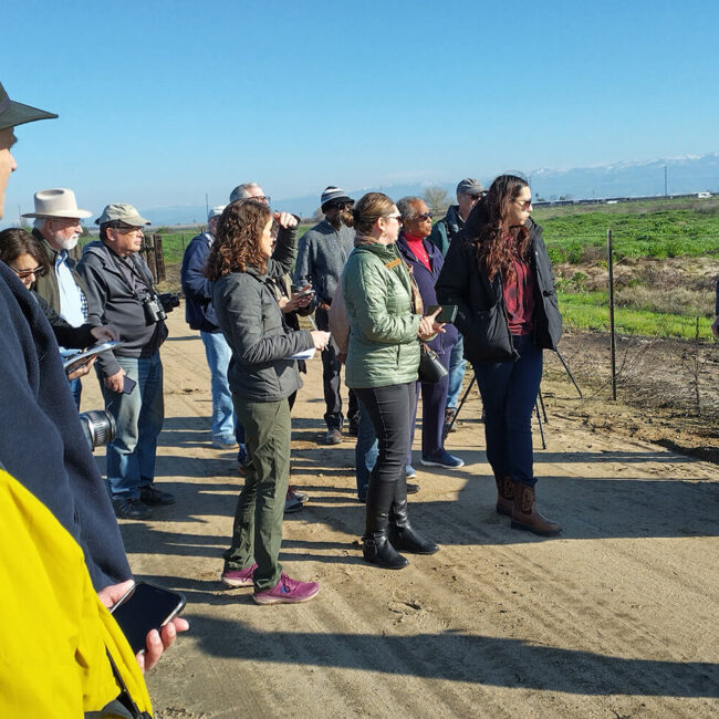 CVJV Board members and guests standing on a dirt road. Afield guide holding a map and describing the restoration project we're visiting.
